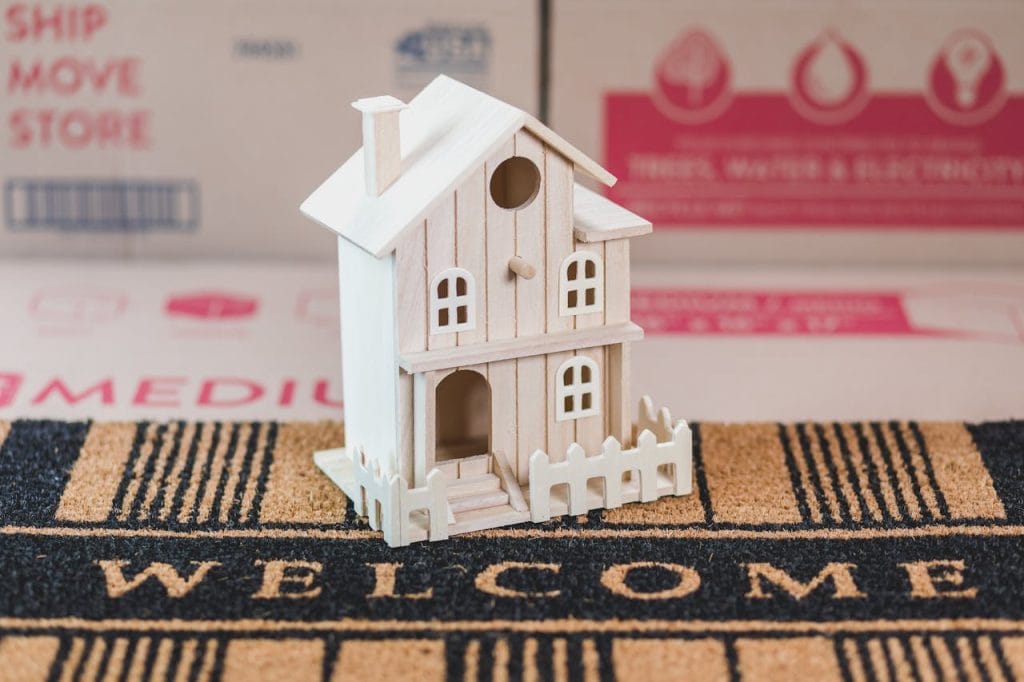 A small wooden house model sits on a welcome mat among moving boxes, symbolizing a new home.