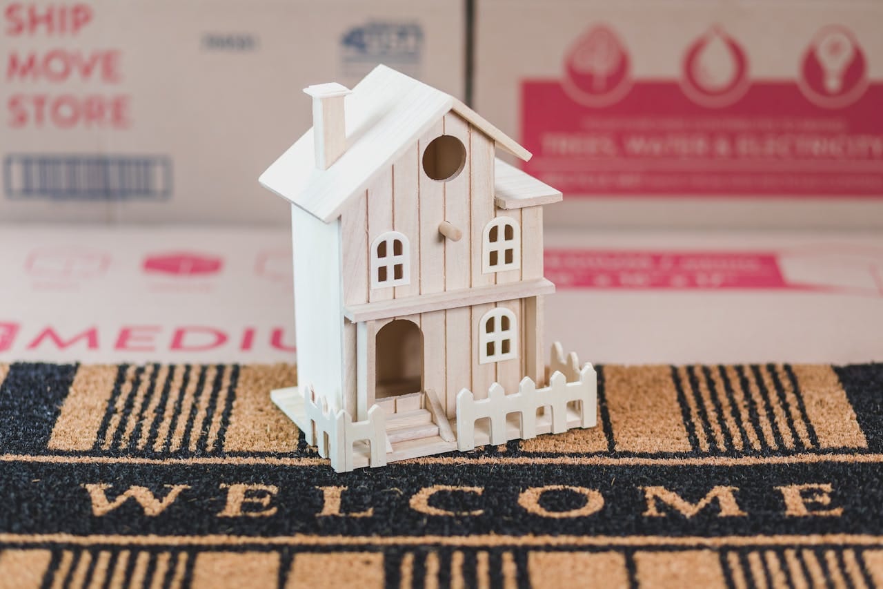 A small wooden house model sits on a welcome mat among moving boxes, symbolizing a new home.